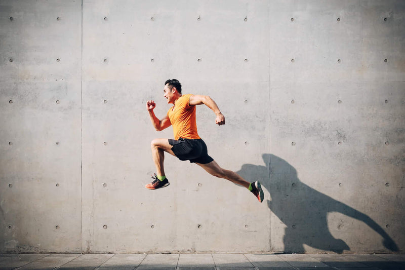 A fit man in an orange athletic shirt and black shorts leaps into the air with a powerful stride. His arms and legs are in motion, showcasing strength and agility. His shadow stretches across the wall behind him, adding a dramatic effect to the image.