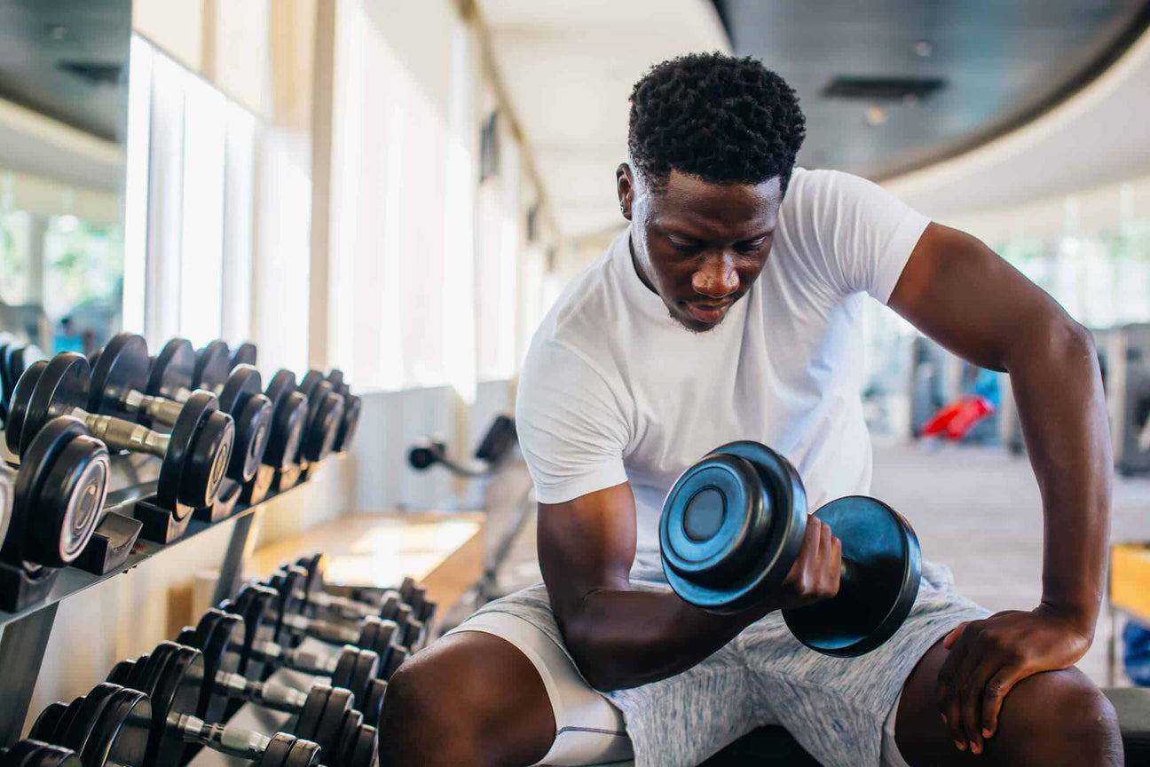 A man in a white t-shirt and gray shorts sits on a bench in a gym, performing a bicep curl with a heavy dumbbell. His focused expression and muscular arms highlight strength training. A rack of dumbbells is visible in the background.