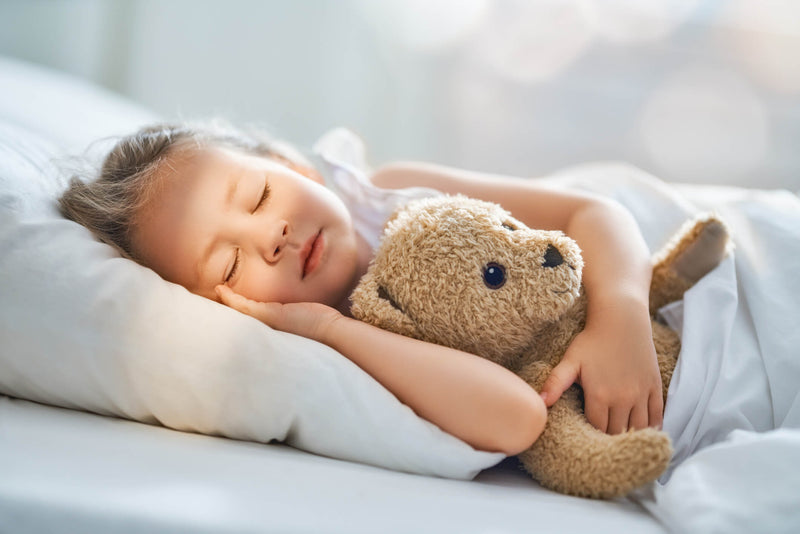 A young child peacefully sleeping in bed, cuddling a plush teddy bear. Sunlight softly filters through the room, casting a warm glow on their relaxed face. The child is tucked under a white blanket, resting on a fluffy pillow.