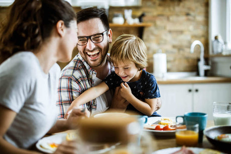 A cheerful family enjoys breakfast together in a bright, rustic kitchen. A father wearing glasses and a plaid shirt laughs while playfully holding his young son, who is reaching toward the mother, who is smiling at them. 