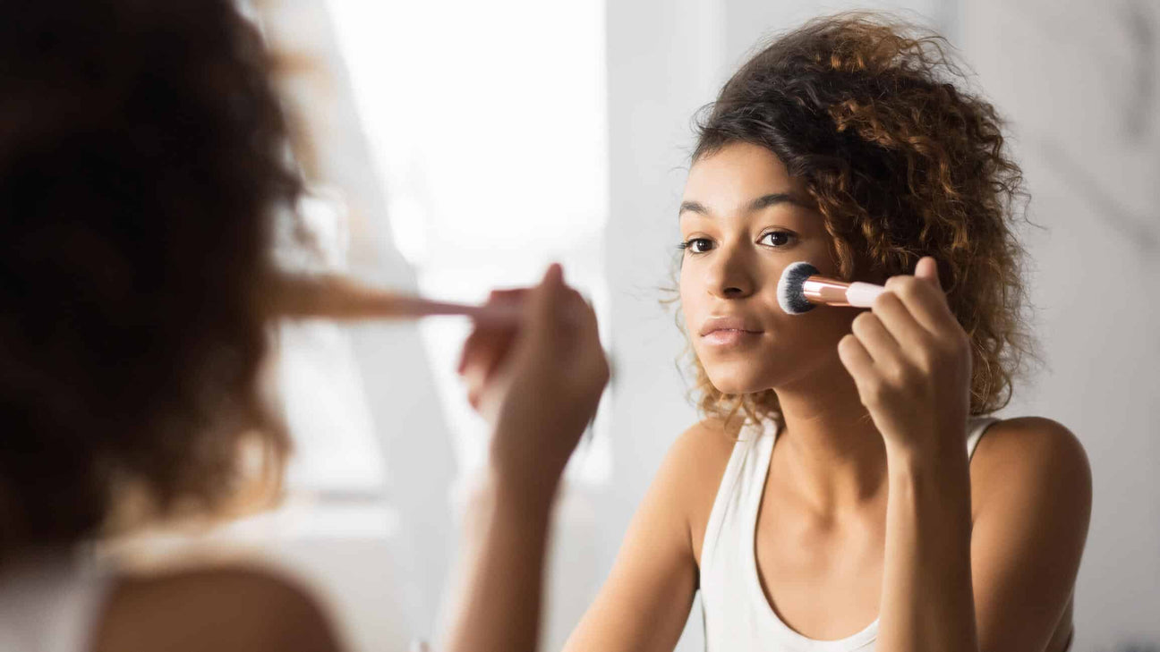 A young woman with curly hair applies makeup with a soft brush while looking into a mirror. Natural light streams in, highlighting her smooth complexion and minimalistic beauty routine. The scene reflects a gentle, natural facial care approach.