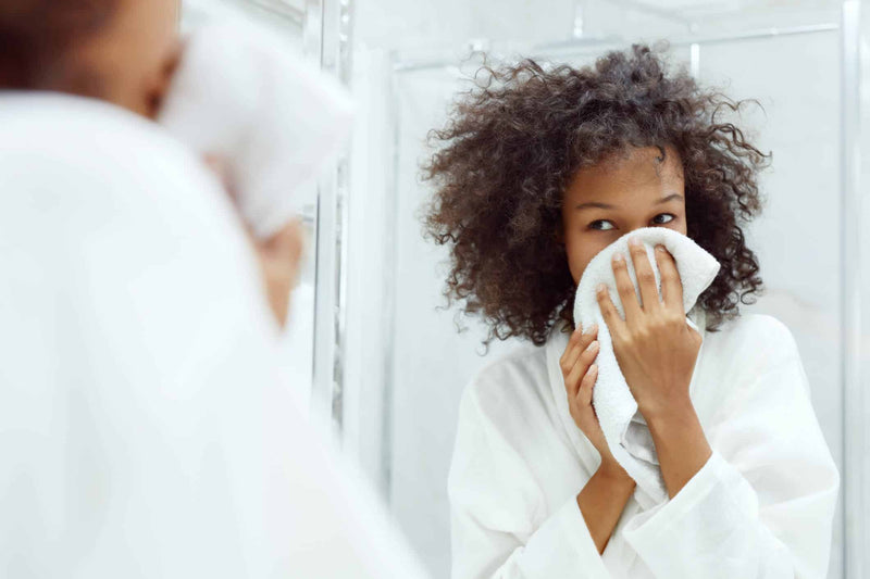 A young woman with curly hair and a white robe gently dries her face with a soft towel in a bright, modern bathroom. Her reflection in the mirror captures her fresh, radiant skin, emphasizing a natural skincare routine. 