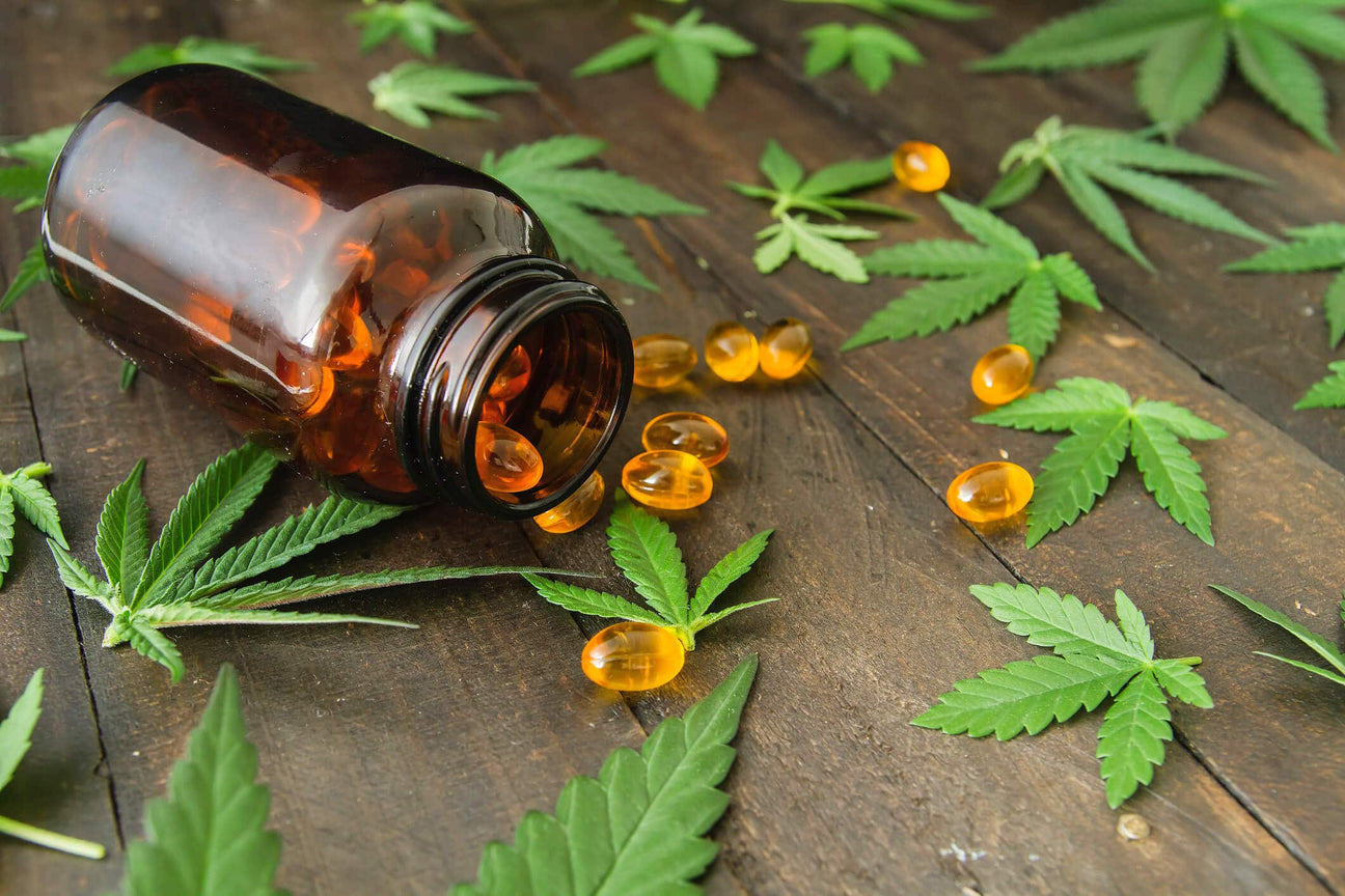 A tipped-over amber glass bottle spills golden CBD capsules onto a rustic wooden surface, surrounded by fresh cannabis leaves. The capsules glisten under the light, emphasizing their smooth, gel-like texture.