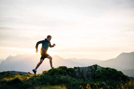 A man in athletic wear runs across a rocky landscape at sunrise, with mountains in the background. The golden light casts a dramatic glow, emphasizing movement, energy, and outdoor adventure. 