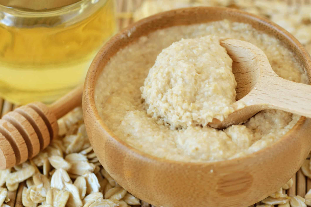 A wooden bowl full of creamy oatmeal and honey scrub, with a wooden spoon scooping out a portion. Surrounding the bowl are scattered oats, a glass jar of golden honey, and a honey dipper, emphasizing the natural ingredients used for body exfoliation.