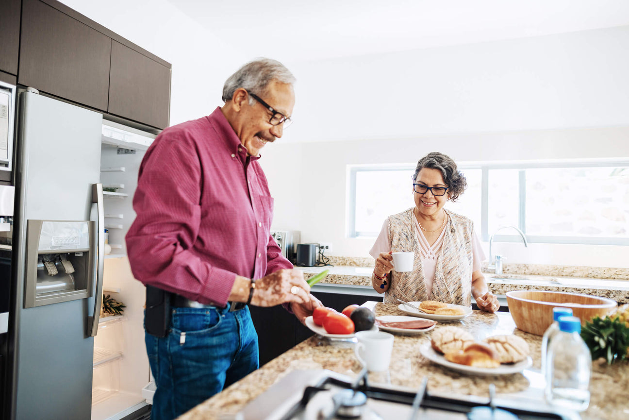 An elderly couple enjoys a morning in their bright, modern kitchen. The man in a maroon shirt and glasses slices fresh vegetables, while the woman in a patterned vest and glasses holds a cup of coffee, smiling warmly. 