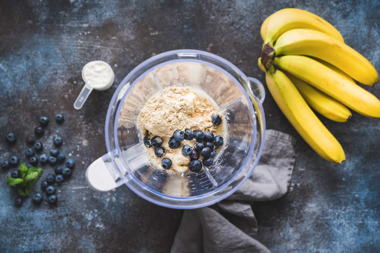 A blender filled with bananas, blueberries, and plant-based protein powder sits on a dark countertop, ready to be blended into a smoothie. A scoop of protein powder, fresh blueberries, and a bunch of ripe bananas are arranged around the blender.