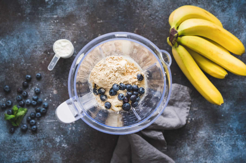 A blender filled with bananas, blueberries, and plant-based protein powder sits on a dark countertop, ready to be blended into a smoothie. A scoop of protein powder, fresh blueberries, and a bunch of ripe bananas are arranged around the blender.