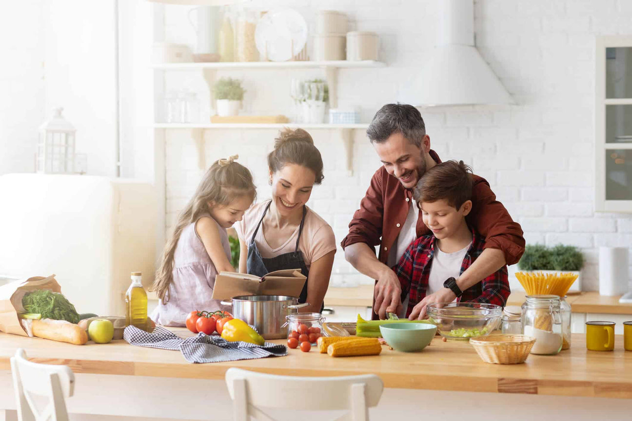 A family prepares a meal together in a bright, modern kitchen. Mother and daughter read a cookbook while father helps his son chop vegetables. The table is filled with fresh produce, pasta, and cooking essentials, creating a warm, lively atmosphere.