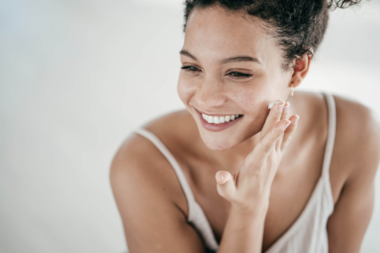 A smiling woman with glowing skin applies a dollop of cream to her cheek, embracing a natural skincare routine. She wears a white camisole. The bright backdrop of the scene conveys self-care, beauty, and healthy skin.