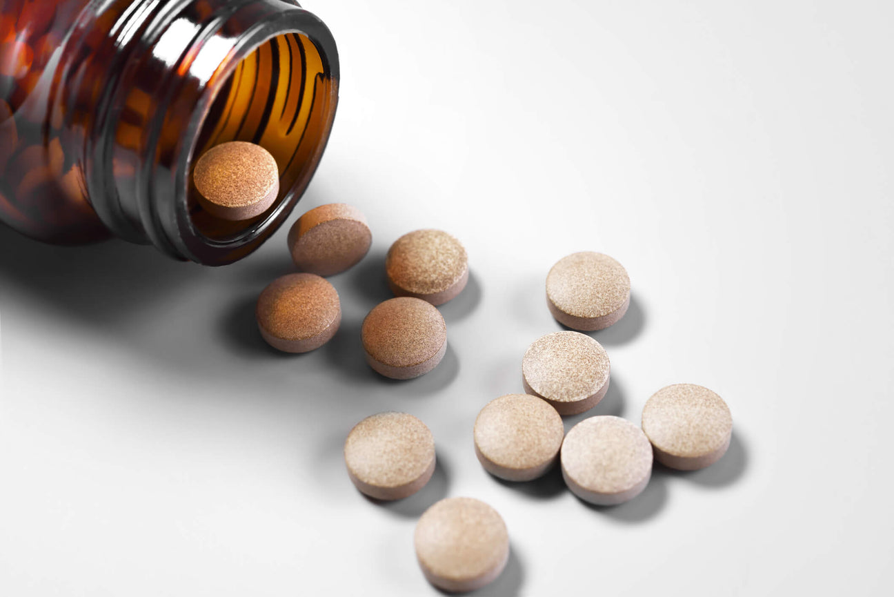 A brown glass bottle tipped over, spilling round brown tablets onto a white surface. The tablets have a slightly textured surface, and the clean background highlights the simplicity of the supplement presentation.