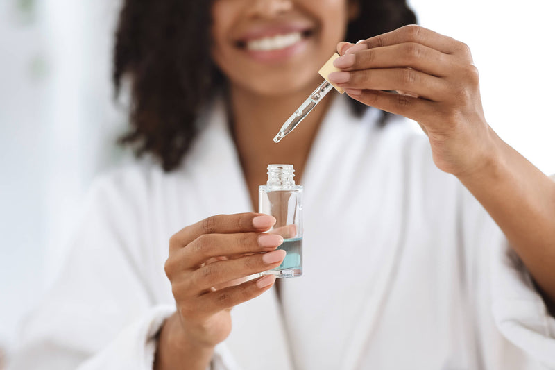 A woman in a white robe smiles as she carefully lifts a dropper filled with a clear blue facial serum from a glass bottle. Her well-manicured nails and glowing complexion highlight a skincare routine focused on hydration and self-care. 