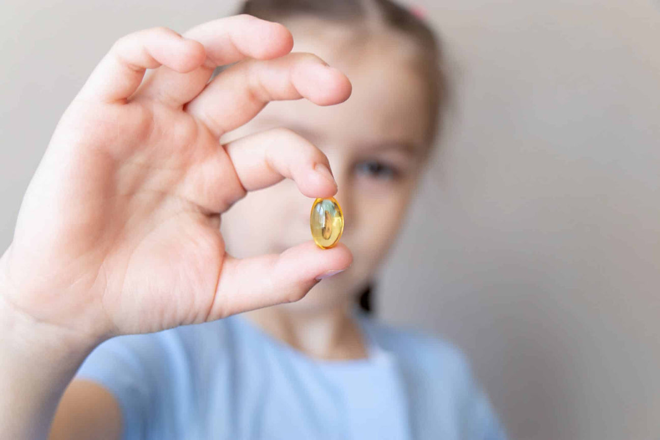 A young child in a light blue shirt holds up a translucent yellow omega-3 supplement capsule between their fingers. The child's face is blurred in the background, focusing attention on the softgel. 