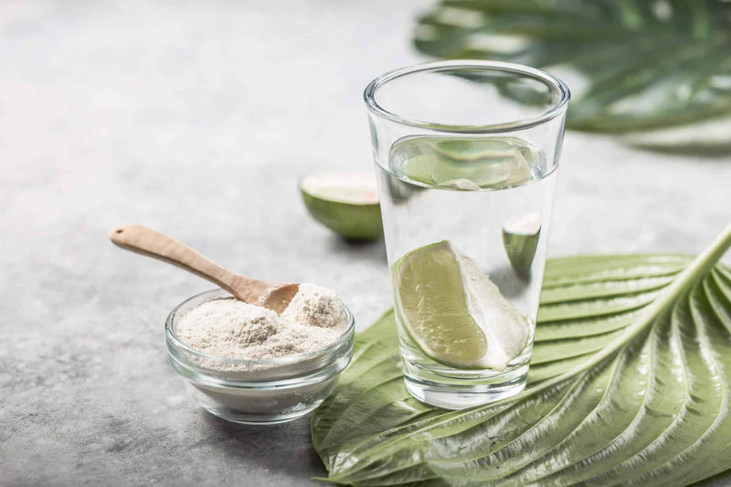 A glass of water with lime slices sits on a green leaf, next to a small glass bowl filled with white collagen powder and a wooden spoon. A halved lime is in the background, emphasizing freshness and wellness.