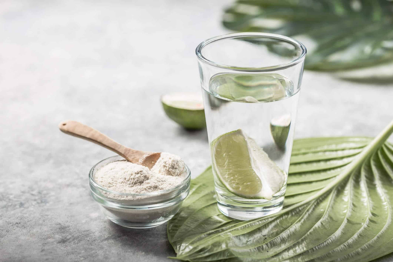 A refreshing glass of water with lime slices sits beside a small bowl of white collagen supplement powder and a wooden spoon. The scene is set on a green leaf with a halved lime in the background, symbolizing natural wellness and beauty support.