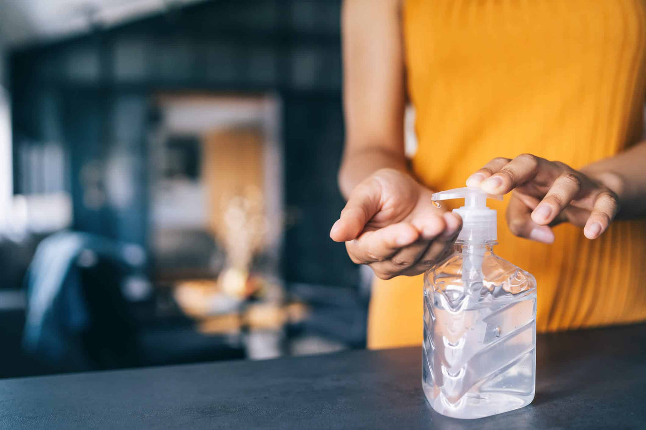 A person in a mustard-colored top dispensing clear hand sanitizer from a pump bottle onto their palm. The background is blurred, showing a cozy indoor setting, emphasizing hygiene and cleanliness.