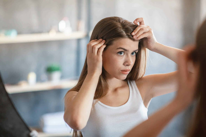 A young woman with long, straight brown hair examines her scalp in the mirror, gently parting her hair with both hands. She has a focused expression.The image conveys themes of hair care, scalp health, and self-care routines.