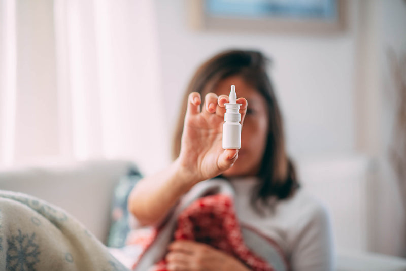 A woman sits on a couch wrapped in a blanket, holding up a small white nasal spray bottle. The background is softly blurred, highlighting the focus on the spray, which appears to be for cold or allergy relief.