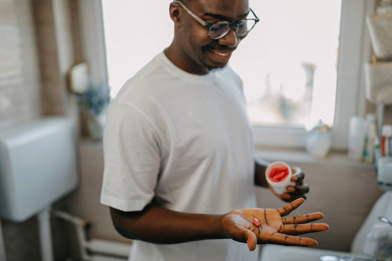 A smiling man wearing glasses and a white t-shirt stands in a bright bathroom, holding a red capsule in his open palm while looking at it. In his other hand, he holds a bottle filled with similar red capsules. 