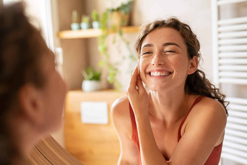 A young woman with curly brown hair and freckles is applying a skincare product to her cheek while looking in the mirror. She is smiling with her eyes slightly squinted, wearing a red tank top. 