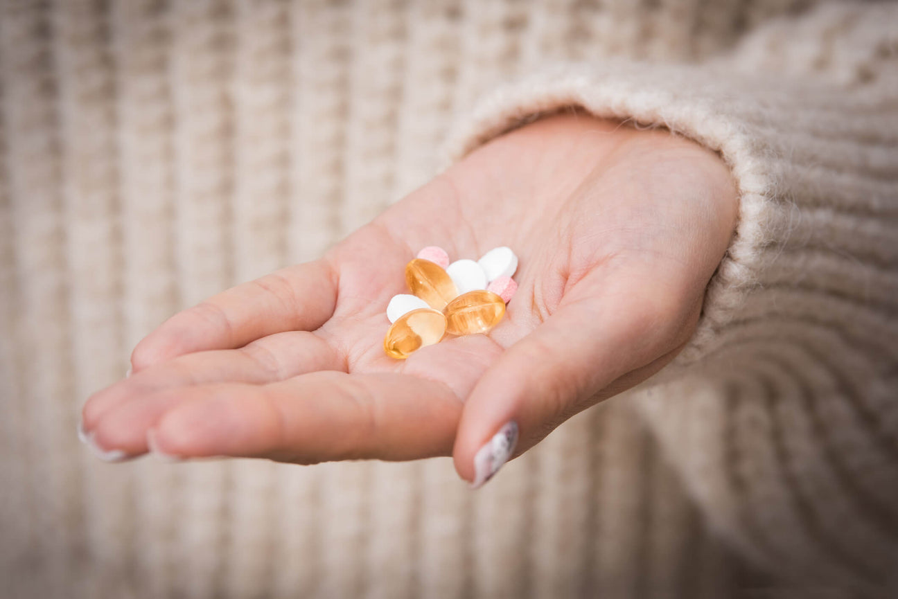 A close-up of a person's hand holding a mix of vitamins and supplements, including soft gel capsules and tablets. The person is wearing a cozy beige knit sweater, and the blurred background adds warmth to the scene.