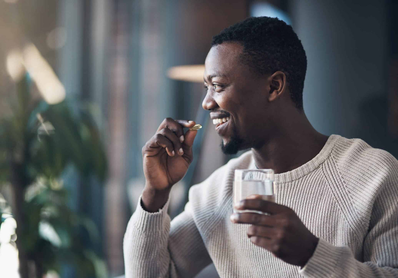 A smiling man in a cozy beige sweater holds a glass of water in one hand and a soft gel supplement in the other, about to take it. The background is softly blurred, with warm natural lighting and indoor greenery.