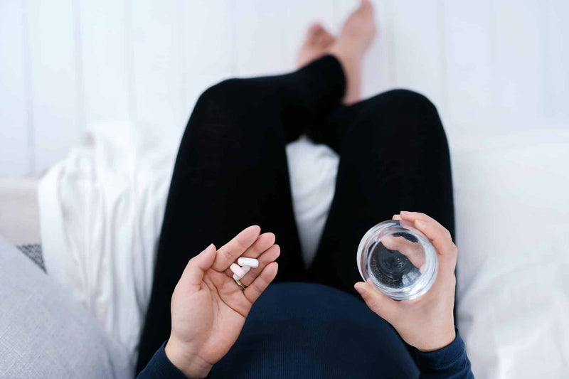 A person sits on a bed wearing black leggings and a dark blue top, holding white capsules in one hand and a glass of water in the other. The soft white bedding and relaxed posture suggest a moment of self-care or supplement intake.