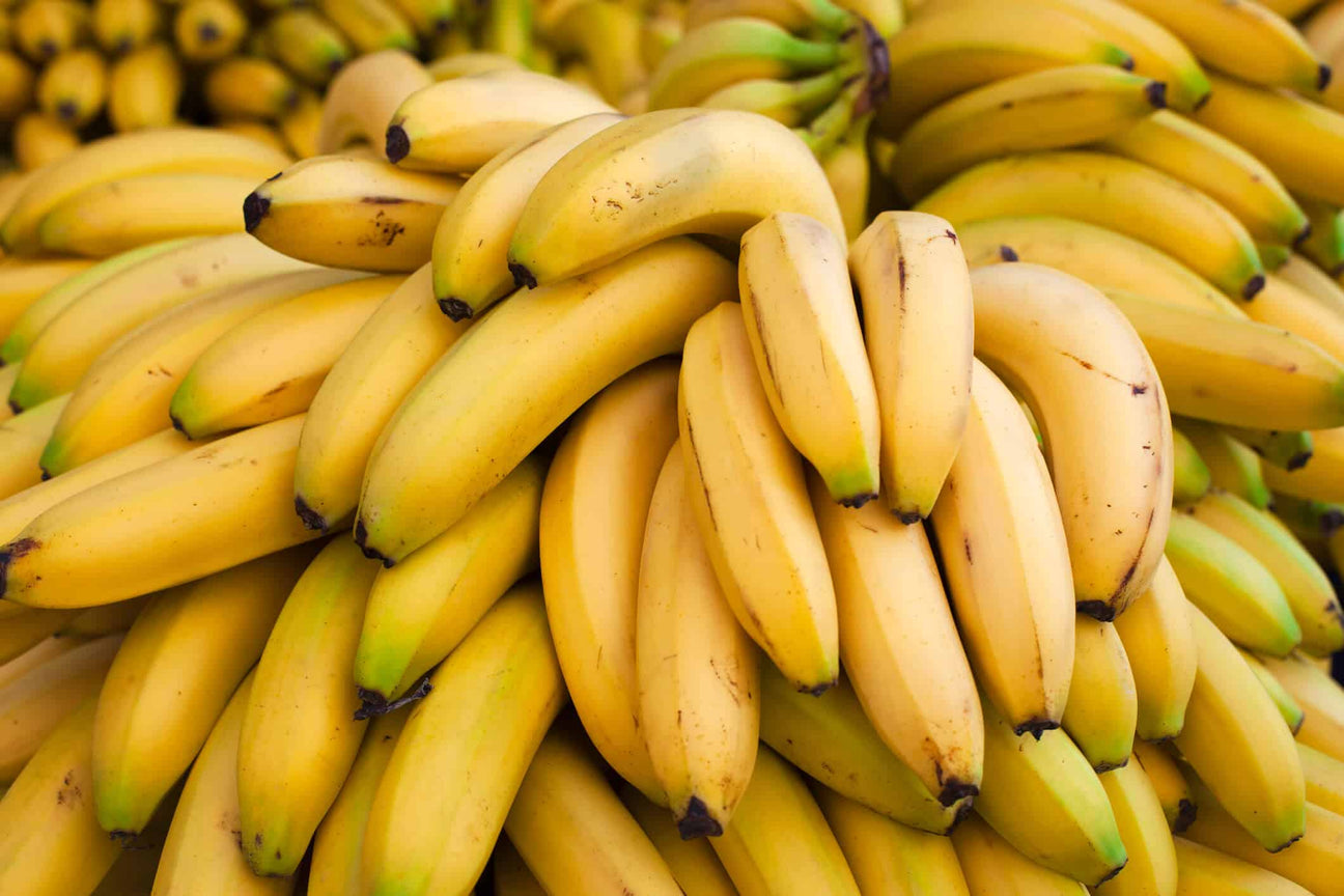 A large pile of ripe yellow bananas with hints of green at the tips. The bananas are stacked closely together, showcasing their smooth texture and natural imperfections.