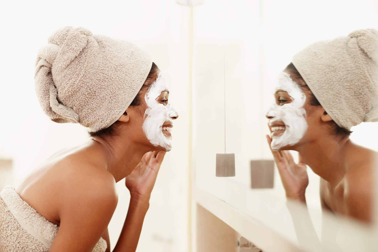 A woman wrapped in a towel with her hair in a matching towel turban smiles at her reflection in the mirror while applying a white facial mask. Her glowing skin and relaxed posture highlight a self-care and skincare routine focused on natural beauty.