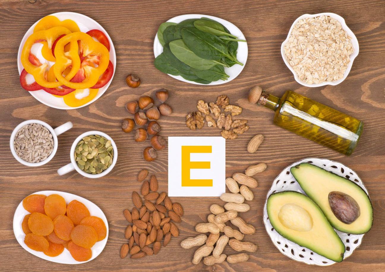 A variety of vitamin E-rich foods arranged on a wooden surface, including avocado, nuts, seeds, spinach, bell peppers, dried apricots, and a bottle of oil. A white card with a bold yellow "E" highlights the nutrient focus.