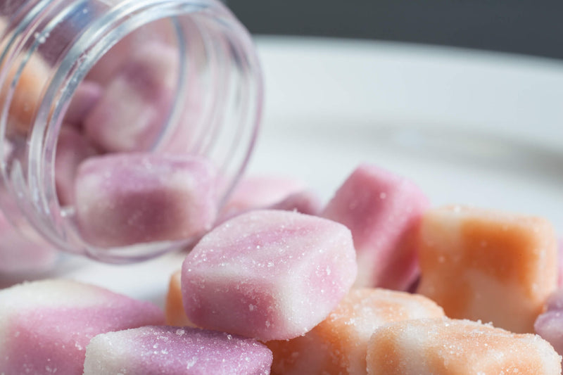 Close-up of colorful CBD gummies spilling out of a clear plastic jar onto a white plate. The gummies are sugar-coated and come in shades of pink, white, and orange.