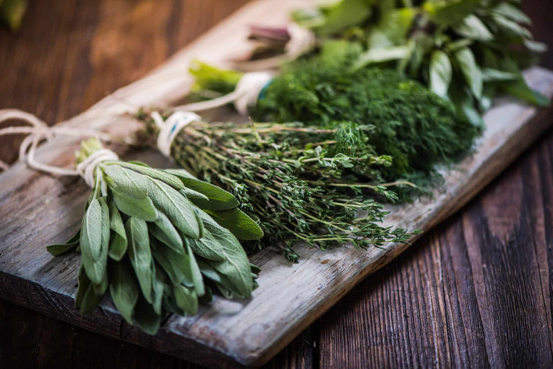 Fresh bundles of herbs, including sage, thyme, dill, and basil, neatly tied with twine, rest on a rustic wooden cutting board. The vibrant green leaves contrast with the warm, dark wood grain. These herbs are used for culinary and medicinal purposes.