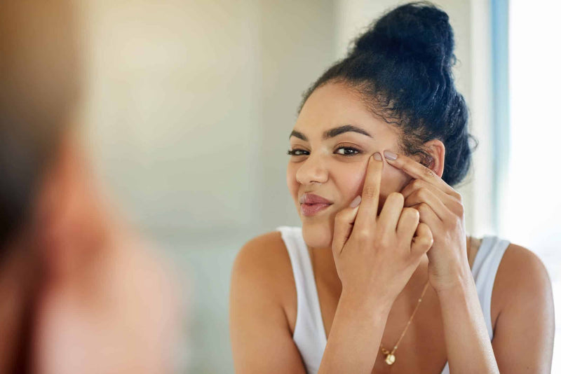 A young woman with curly hair tied in a bun is standing in front of a bathroom mirror, gently squeezing a blemish on her cheek. She wears a determined expression on her face and a white tank top with a delicate gold necklace. 