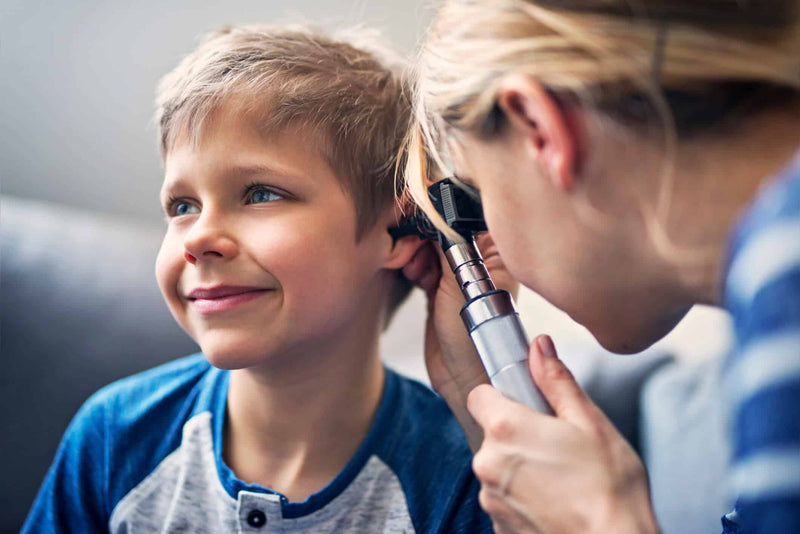 A young boy with short blonde hair and a blue raglan shirt smiles while a woman, possibly a doctor or parent, examines his ear with an otoscope. The woman, whose face is partially visible, leans in closely to check his ear health.