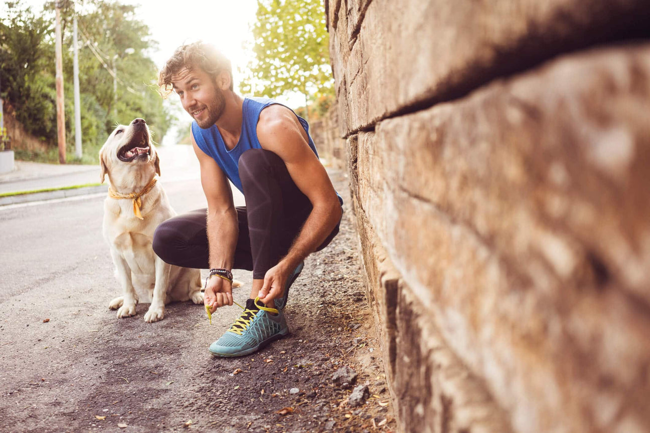 A fit man wearing a blue sleeveless shirt and black leggings crouches down on a paved road to tie his bright turquoise running shoes. A happy Labrador Retriever wearing a yellow bandana sits beside him, looking up at him with its mouth open. 