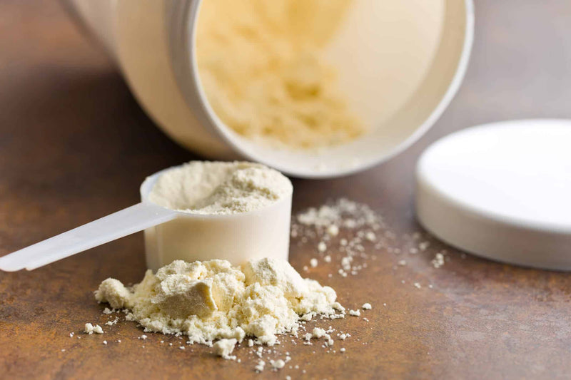 A scoop of protein powder sits on a wooden surface, with an open container in the background. Some powder is spilled around the scoop, emphasizing its fine texture. The image highlights nutrition and fitness supplements.