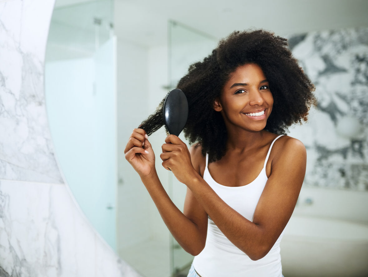 A smiling woman with voluminous, curly hair brushes her strands in a bright, modern bathroom. She wears a white tank top and stands in front of a large mirror. Her healthy, radiant hair highlights the benefits of proper hair, skin, and nail care.