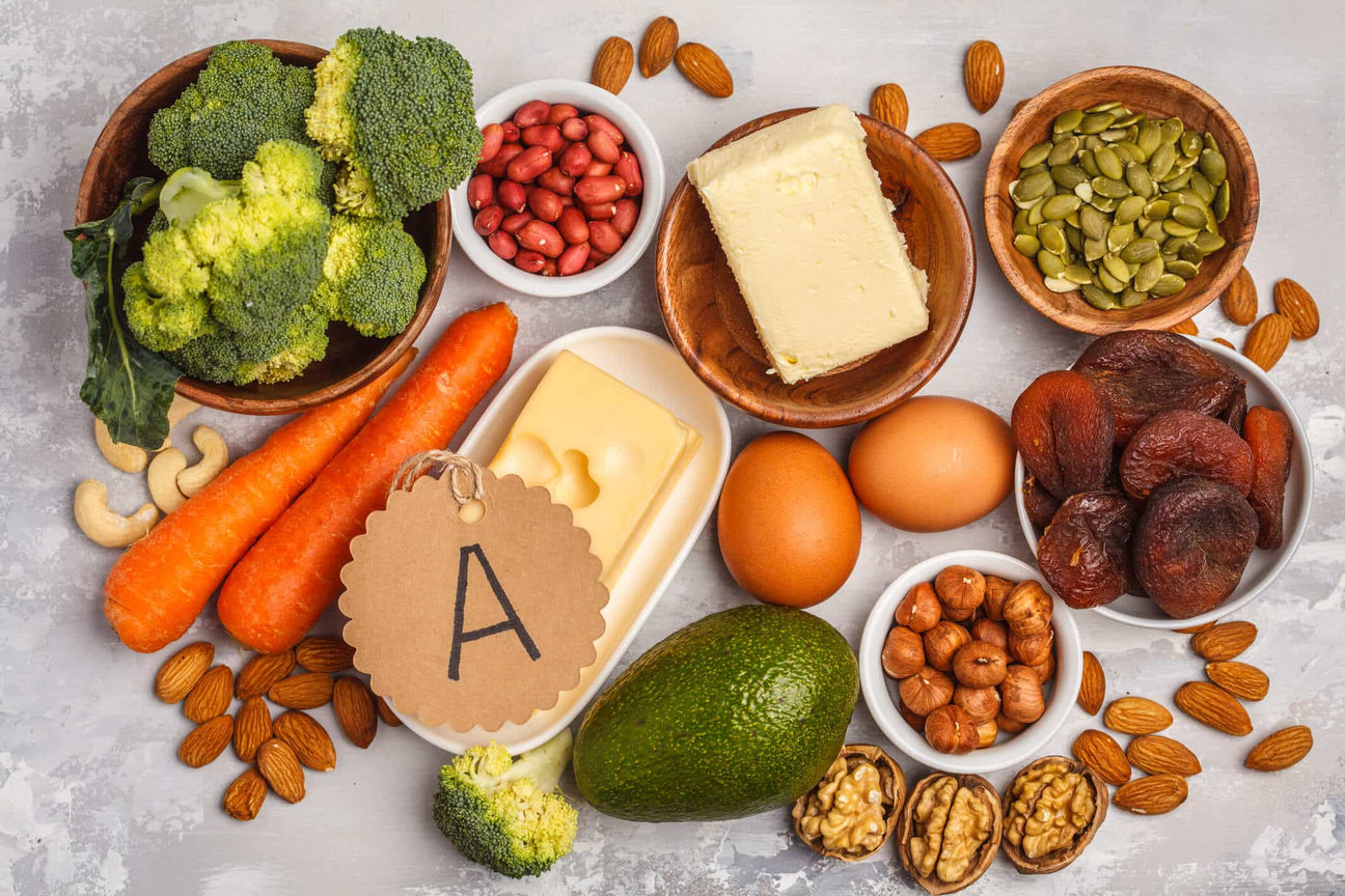 A variety of vitamin A-rich foods arranged on a textured surface, including carrots, broccoli, eggs, butter, cheese, dried apricots, nuts, and seeds. A small tag labeled "A" highlights the nutrient focus.