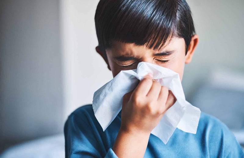 A young boy with dark hair and a blue long-sleeve shirt tightly squeezes his eyes shut as he blows his nose into a tissue. His expression suggests discomfort, possibly from a cold or allergies.