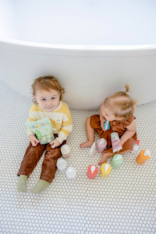 Two young children sitting on a white tiled bathroom floor near a bathtub, surrounded by colorful bottles of bath and skincare products. 