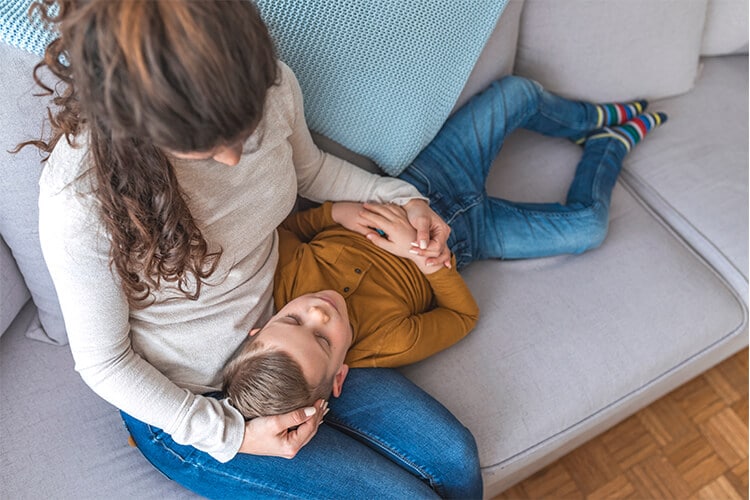 A mother sits on a light gray couch, gently cradling her young child’s head in her lap. The child, dressed in a mustard-colored top and blue jeans, lays down with their eyes closed, wearing colorful striped socks.