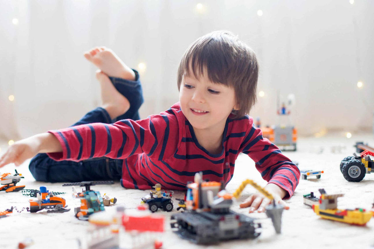 A young child with shaggy brown hair and a red striped shirt smiles while playing with colorful building blocks and toy vehicles on a soft white rug. Warm fairy lights twinkle in the background, creating a cozy and playful atmosphere.