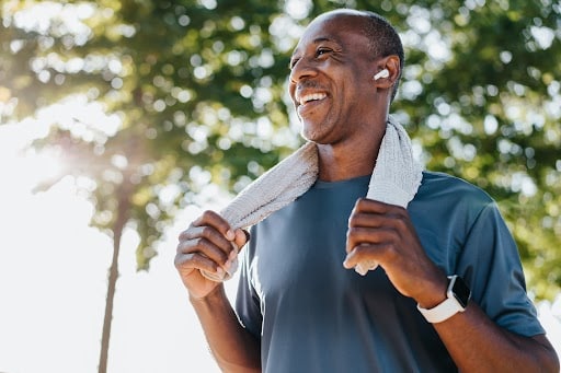 Smiling man in athletic attire standing outdoors, holding a towel around his neck. He is wearing wireless earbuds and a fitness watch, with sunlight filtering through a backdrop of green trees, suggesting a post-workout moment.