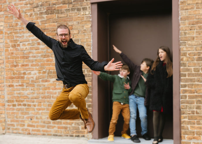Tim OBrien, owner of The Healthy Place, leaping in the air with a wide smile and his arms outstretched. Behind him are several of his children in a doorway posing and laughing. 
