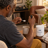 Vital Nutrients Glycine Powder container on a table as an older man adds a spoonful of powder to a mug