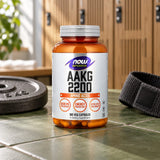 NOW Sports AAKG 2200 bottle on a tiled floor beside a weight plate and a resistance band, lockers behind