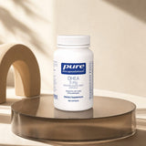 Pure Encapsulations DHEA 5 mg bottle on a bathroom counter beside a morning supplement organizer