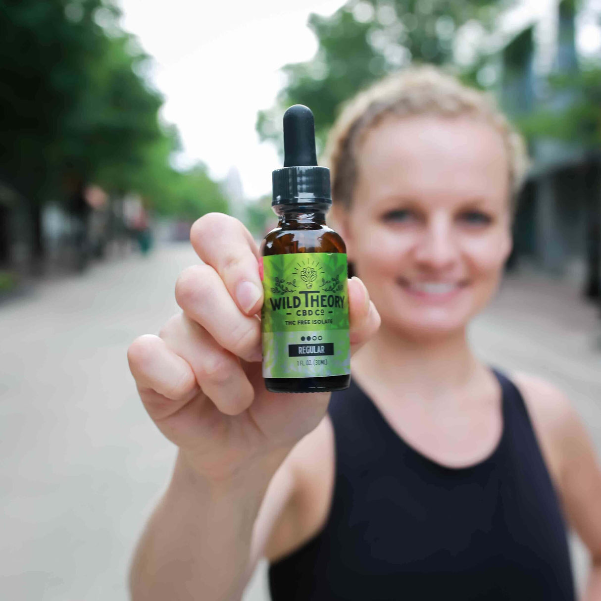 A woman holding a bottle of Wild Theory CBD oil with a green label, smiling in the background on a city street.