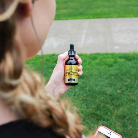 A person holding a bottle of Wild Theory CBD Co. Full Spectrum CBD Oil, labeled "Maximum Strength" with a yellow and green design. They are standing outdoors on a grassy area, with a smartphone visible in their other hand.