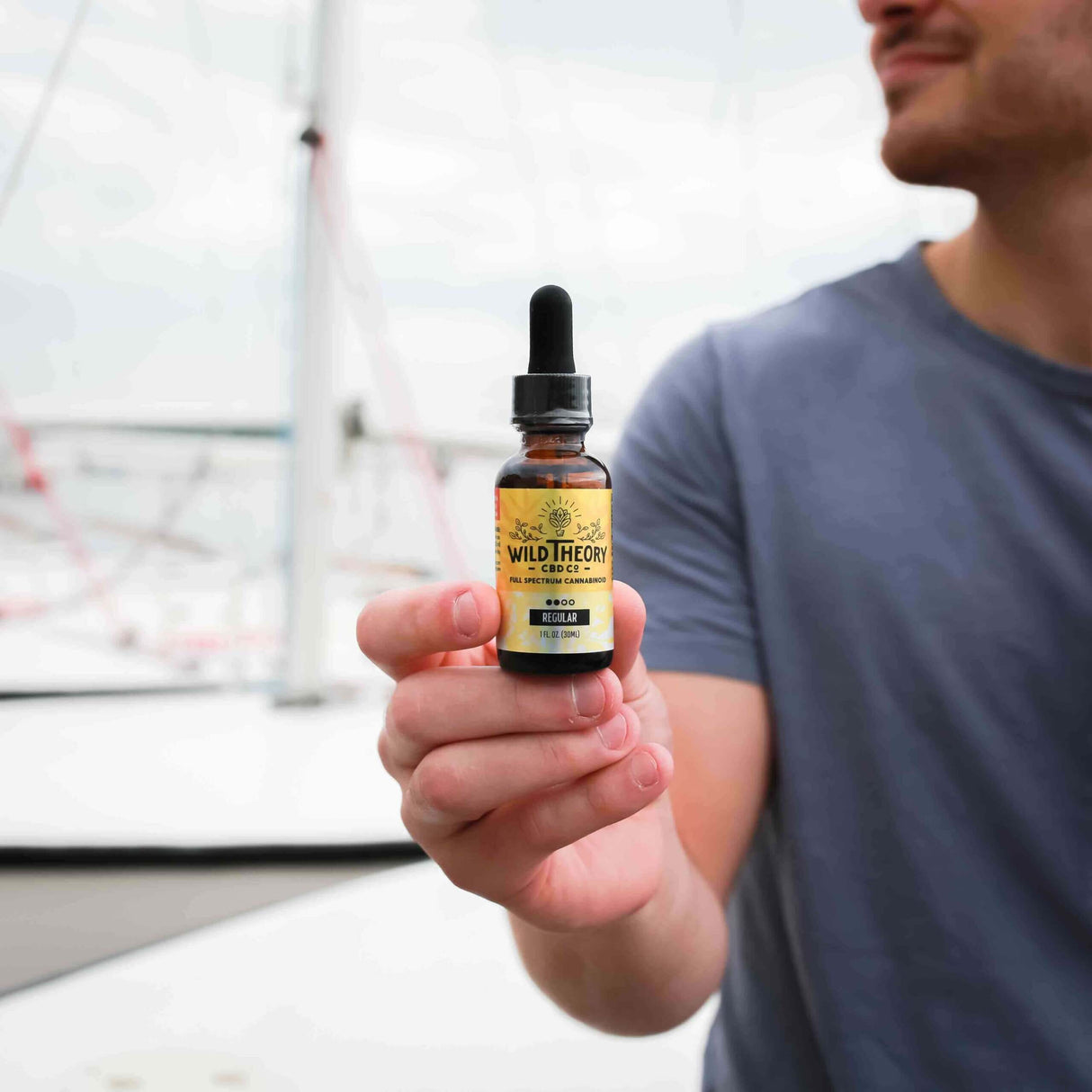 A close-up of a man holding a bottle of Wild Theory CBD oil, labeled "Full-Spectrum Cannabinoid," with a boat or marina in the background.